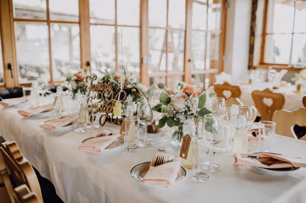Liebevoll gedeckte Hochzeitstafel mit Rosen und Holzdekoration vor großen Fenstern